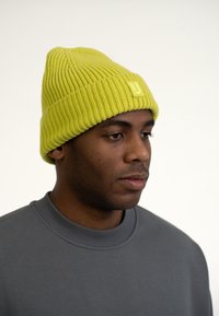 Gorro de punto amarillo con textura acanalada y un pequeño parche con logo. Llevado con un sudadera gris con cuello redondo.
