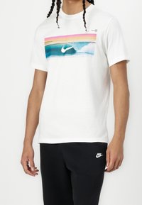 Bílá bavlněná tričko s barevnou vlnou grafikou v odstínech růžové, modré a bílé, s logem Nike uprostřed.