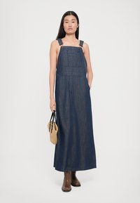 Rochie din denim cu bretele și buzunare frontale, având o nuanță albastră și o siluetă lungă, dreaptă. Modelul ține o geantă împletită.