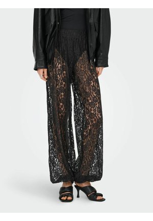 Personne portant un pantalon large en dentelle noire avec un short noir en dessous, une veste en cuir noire et des sandales à talons noires.