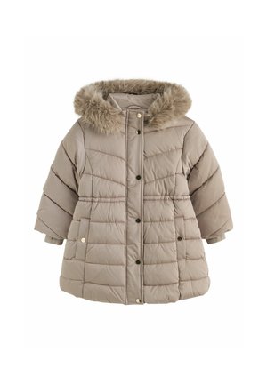 Ung pige iført en beige dunjakke med pelsfor, blå cardigan, denim nederdel og brune støvler stående mod en ensfarvet væg.