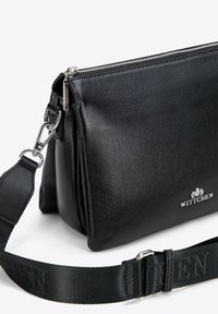 Czarna skórzana torebka na ramię z teksturowanym wykończeniem, wyposażona w srebrny zamek błyskawiczny, regulowany pasek i wytłoczone logo na froncie.