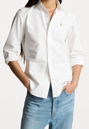 Camicia bianca button-up in cotone, con maniche lunghe, colletto e un piccolo logo ricamato sul petto. Abbinata a un paio di jeans blu chiaro.