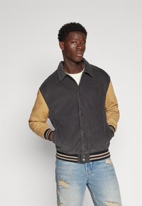 Veste en velours côtelé gris foncé avec des manches beige, dotée de poignets et d'un ourlet côtelés, et fermeture à boutons. Portée avec un jean bleu déchiré.