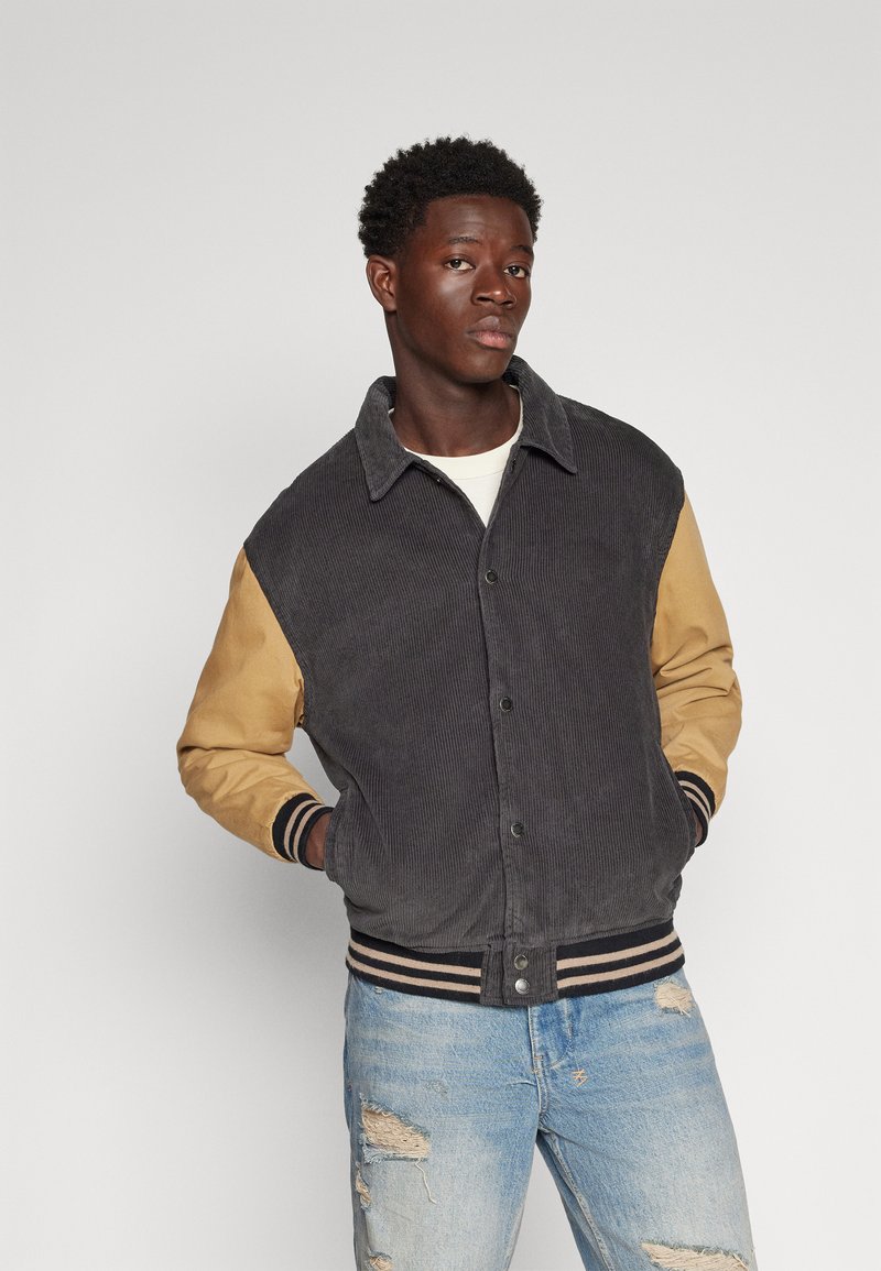 Veste en velours côtelé gris foncé avec des manches beige, dotée de poignets et d'un ourlet côtelés, et fermeture à boutons. Portée avec un jean bleu déchiré.