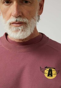 Sudadera burdeos con un cuello redondo acanalado, que presenta un logotipo negro y amarillo con alas en el lado izquierdo. Textura suave.