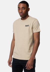 Béžové bavlnené tričko s krátkymi rukávmi, okrúhlym výstrihom a malým čiernym logom Lonsdale na ľavej hrudi. Model má na sebe čierne nohavice.