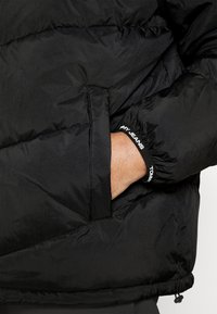 Černá péřová bunda s prošívaným vzorem, s boční kapsou a elastickými manžetami s logem "TOMMY JEANS". Hladká, lesklá textura.