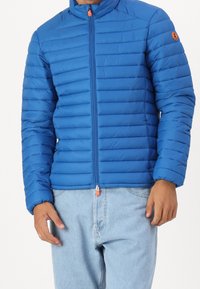 Blauwe pufferjas met horizontale stiksels, hoge kraag, ritsluiting en oranje ritstrekkertje. Gemaakt met lichtblauwe jeans.