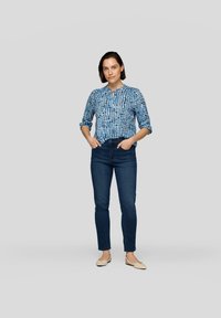 Blau gemustertes Hemd mit Knöpfen und langen Ärmeln, kombiniert mit dunklen, engen Jeans und hellbeigen flachen Schuhen.