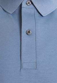 Ljusblå polo skjorta gjord av texturerad tyg, med en tre-knappars knäppning och mjuk krage. Anmärkningsvärd sömdekoration nära knapparna.