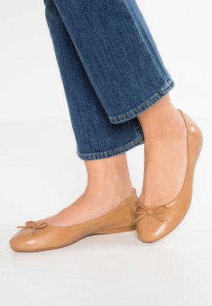 Brune læder ballethæle med rund tå og en lille sløjfedetalje foran. Glat tekstur og lave, flade såler. Jeans synlige ovenfor.