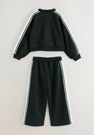 Černý sportovní tracksuit obsahující zkrácenou bundu a kalhoty širokého střihu. Bílé boční pruhy zdobí rukávy a strany kalhot.