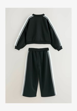 Črna atletska trenirka, ki vključuje krajšo jakno in široke hlače. Bele stranske črte krasijo rokave in stranice hlač.