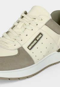 Baskets Michael Kors beige et taupe avec lacets à l'avant et panneau latéral perforé sur semelle blanche.