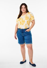 Camiseta blanca con estampado amarillo de flores, combinada con pantalones cortos de mezclilla azul con dobladillos enrollados. La modelo lleva unas bailarinas azul marino.