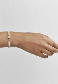 Pulsera de plata con pequeñas perlas blancas. Presenta un diseño de cierre sencillo y texturas suaves. Se lleva en una mano sobre un fondo liso.