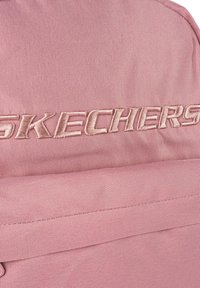 Mochila de tela rosa con una superficie texturizada y un prominente logo bordado de "SKECHERS" en un tono más claro. Incluye detalles como un bolsillo frontal.