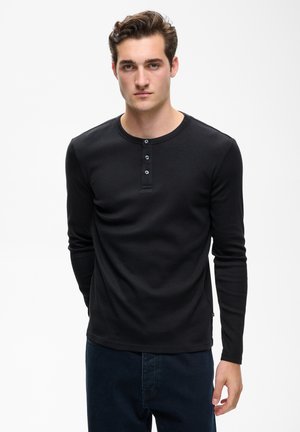 Giovane uomo con capelli corti e mossi che indossa una maglia henley nera a maniche lunghe e pantaloni scuri, in piedi davanti a uno sfondo bianco semplice.