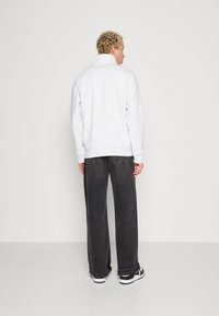 Hvid fleece-pullover med høj krave, parret med løstsiddende mørke denim jeans. Modellen bærer sorte og hvide sneakers.