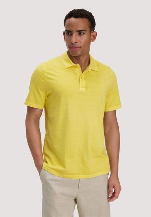 Man die een felgeel poloshirt met korte mouwen en lichtbeige broek draagt, staat met één hand in zijn zak en kijkt opzij.