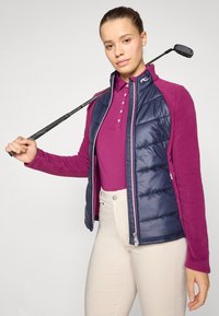Tamnoplavi jakni prsluk preko ljubičaste majice s dugim rukavima, uparen s svijetlosmešavim hlačama, držeći golf palicu.
