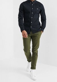 Mörkblå knapp-up skjorta med vita knappar, i kombination med olivgröna slim-fit byxor och vita sneakers, med en liten logotyp på skjortan.