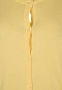 Primo piano di un cardigan giallo abbottonato con un bottone mancante, che mostra una fessura nel tessuto.