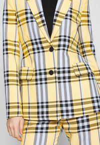 Blazer à carreaux jaune, blanc, noir et gris avec des boutons noirs, associé à un pantalon à carreaux assorti, porté sur un top noir.