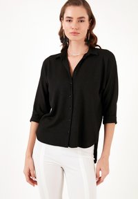 Schwarzes Button-Down-Hemd aus strukturiertem Stoff, mit hochgerollten Ärmeln und lockerer Passform; getragen mit weißen Hosen mit seitlichen Streifen.