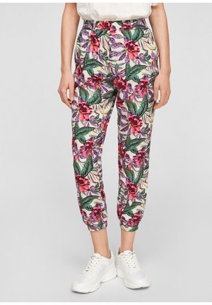 Femme portant des baskets blanches et un pantalon taille haute à fleurs avec des feuilles vertes et des fleurs roses, debout devant un fond gris clair.