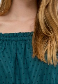 Zbliżenie na szyję i ramię kobiety z długimi blond włosami, ubraną w turkusową, teksturowaną bluzkę bez ramiączek.