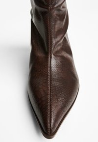 Cizmă din piele maro, cu glezna, cu vârf ascuțit, finisaj texturat și cusături vizibile pe margine. Design simplu, fără accesorii.