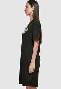 Juodas medvilnės t-shirt suknelė, pasižyminti laisvu siluetu, trumpomis rankovėmis ir šoninėmis plyšėmis. Priekyje yra drąsus baltas užrašas.