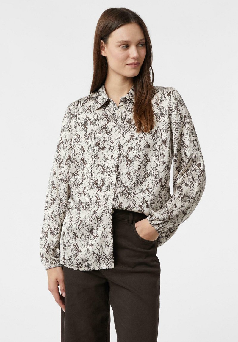 Woman with long brown hair wearing a loose-fitting, long-sleeve snake print blouse and dark pants, standing with one hand in pocket.