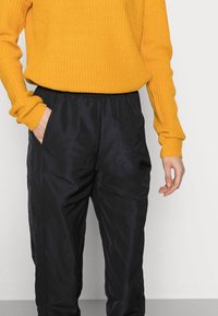 Oranžový pletený svetr s ribbovanou texturou; černé kalhoty s elastickým pasem a hladkým finišem; ruka v kapse; minimalistické pozadí.