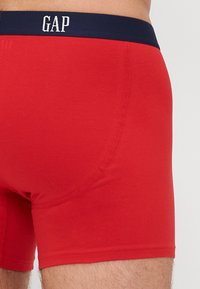 Boxeri din bumbac roșu cu un elastic navy ce prezintă inscripția "GAP" în litere albe. Țesătura pare netedă, având un design ajustat.