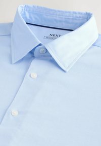 Ljusblå button-up skjorta i texturerad tyg, med vikbar krage och vita knappar. Märkesetikett synlig inuti kragen.