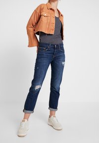 Jeansjack in roestkleur, cropped ontwerp met twee borstzakken. Gestreept tanktop, donkerblauwe distressed jeans met opgerolde mouwen, en witte sneakers.