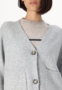 Šedý vlněný cardigan s výstřihem do V, dvěma předními kapsami a dvěma velkými knoflíky. Pod ním je vidět béžový svetr s černým pruhem.