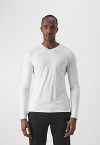 T-shirt de sport à manches longues blanc avec un design à col en V. Fabriqué en tissu lisse, il présente un logo subtil sur la poitrine et une coupe ajustée.