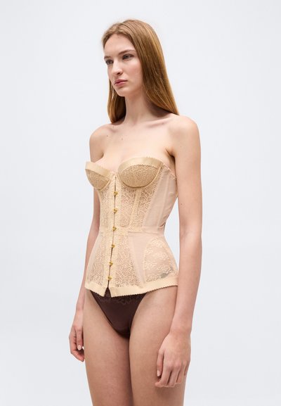 Νεαρή γυναίκα που φοράει μπεζ κορσέ από δαντέλα με χρυσά κουμπιά και σκούρα καφέ εσώρουχα από δαντέλα, στέκεται μπροστά σε απλό ανοιχτό φόντο.