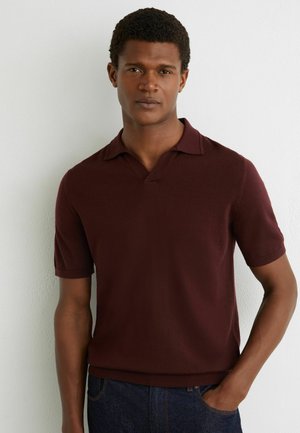 Egy férfi áll fehér fal előtt, rövid ujjú sötét bordó galléros pólót és sötét farmert visel, egyik kezét zsebében tartva.