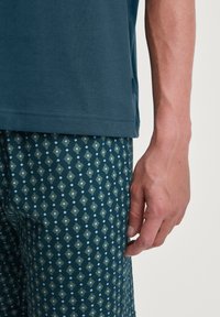 Chemise à manches courtes teal foncé associée à un pantalon vert à motifs présentant un design géométrique en bleu clair et beige. Texture douce du tissu visible.