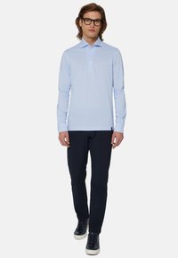 Svetlo modra polo majica z dolgimi rokavi in ovratnikom, opremljena s tremi gumbi, v kombinaciji s temnimi hlačami in športnimi supergami. Gladka tekstura.