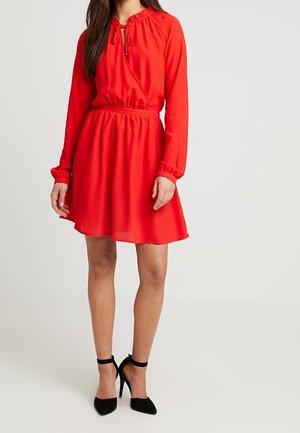 Vrouw draagt een knielange rode jurk met lange mouwen en een geplooide taille, gecombineerd met zwarte hoge hakken met enkelbandje, staand voor een effen achtergrond.