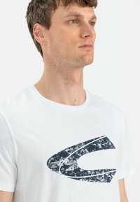 Hvid bomulds-T-shirt med et stort, struktureret marineblåt logo på forsiden. Rund halsudskæring og korte ærmer med en afslappet pasform.