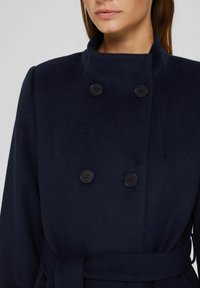 Marineblauwe wollen blended jas met een dubbelzijdig ontwerp, voorzien van zwarte knopen, een hoge kraag en een stoffen riem in de taille.