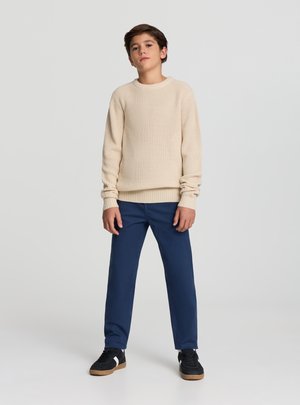 Maglione color beige a coste con maniche lunghe, abbinato a pantaloni blu scuro. Il completo è completato da scarpe da ginnastica nere con suole marrone chiaro.