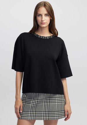 Maglione oversize nero con maniche corte, caratterizzato da un colletto decorato. Abbinato a una gonna a quadri bianchi e neri.